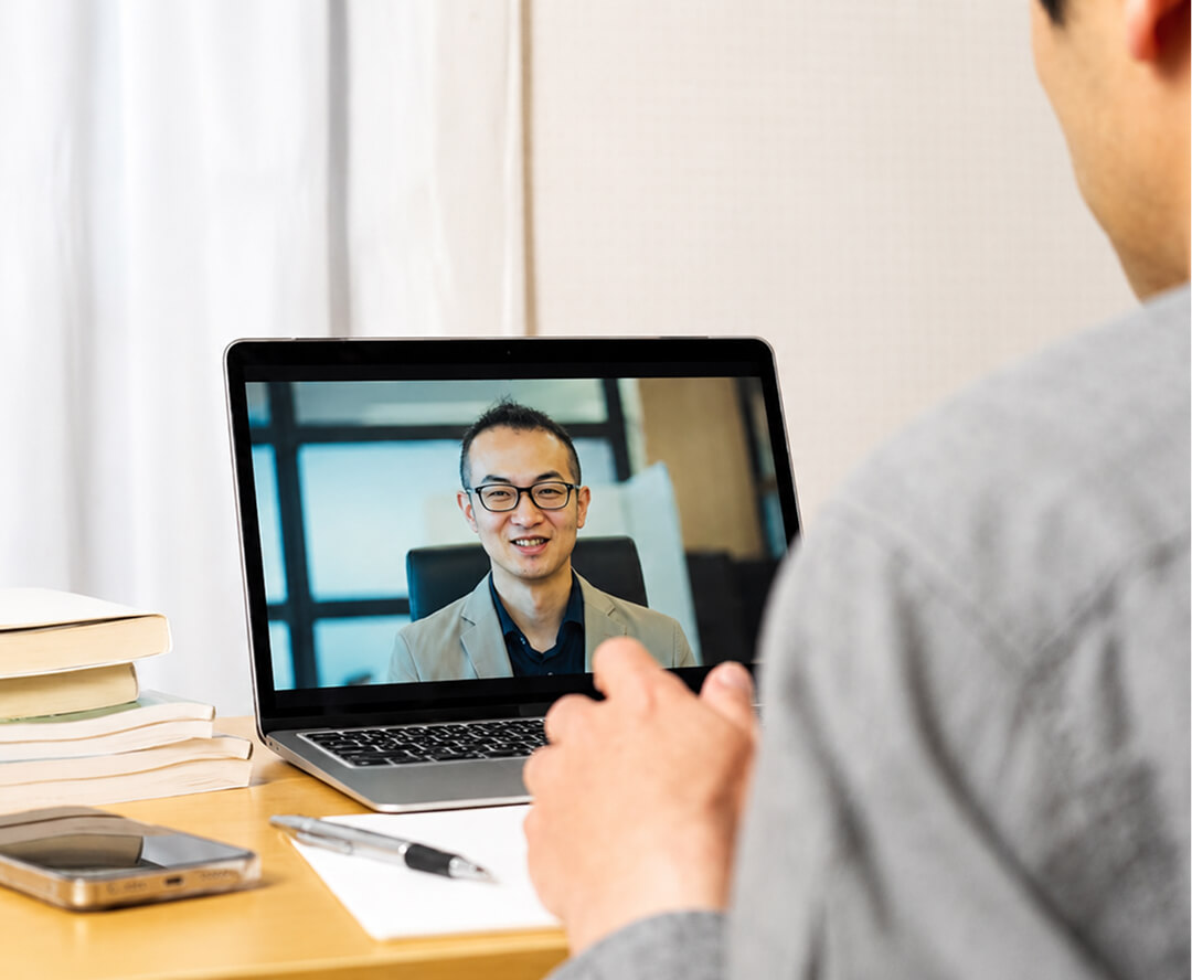 机に座る人物が、ノートパソコンの画面に映る笑顔の男性スタッフとオンラインで会話している様子