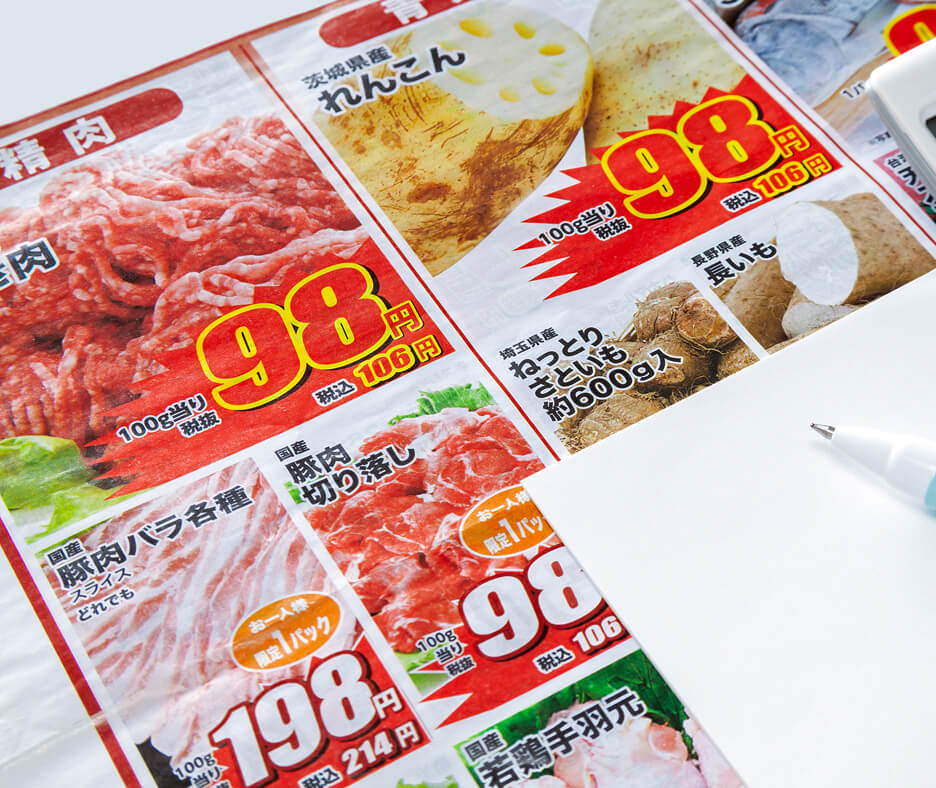精肉や野菜の写真に赤い値札が並ぶスーパーの特売チラシと、横に白い紙とペンが置かれている